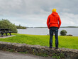 © mark_gusev - A man in a red jacket is looking at a lake lake standing on a stone fence. The sky is cloudy and the water is calm. Travel and tourism. Calm and relaxing mood.
