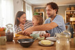 © Stockphotodirectors - Parents and child gather around a table for breakfast, sharing laughter while preparing food. The warm and cozy atmosphere enhances their quality family time.