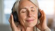 © ЮРИЙ ПОЗДНИКОВ - Senior woman with gray hair enjoys music wearing headphones, eyes closed, in a bright indoor setting with soft lighting and blurred background