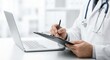 © guobao - Professional doctor in white coat writing medical notes on clipboard at hospital desk with laptop and stethoscope for healthcare concept