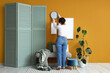 © Pixel-Shot - Young woman hanging clock on orange wall in room, back view