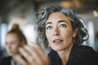 © EricMiguel - Mature female executive explaining in a meeting — close - up of gray curly hair, freckles, gold hoop earring, dotted blazer, upward gaze and expressive hand