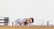 © Prostock-studio - A man is sleeping on his desk in an office setting. He has his head resting on his arm and is surrounded by folders, a coffee mug, and a laptop. The workplace looks quiet.