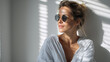 © GrayFrog - Stylish woman with round sunglasses and casual shirt standing in soft sunlight with window shadow on wall.