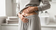 © julijadmi - Woman in gray loungewear holds her stomach while standing in a bright bathroom with towels and a sink visible in the background