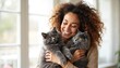 © miss irine - Smiling woman with curly hair holds two large fluffy gray Maine Coon cats. Happy owner cuddles her pets indoors near a window. Joyful connection between human and feline friends.