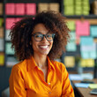 © miss irine - Smiling woman with glasses in orange shirt. She has curly hair and works in a colorful office. This businesswoman is happy and confident at her desk, looking forward to success.