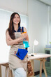 © MINAE - Young woman smiling, wearing an apron, and holding cleaning supplies, symbolizing dedication to home maintenance and professional housekeeping services