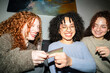 © Studio Marmellata - Three young women with curly hair laugh together as one holds out a credit card, suggesting a shared purchase or transaction.