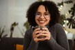 © Studio Marmellata - A young woman with curly hair and glasses smiles while holding a mug of a warm beverage.