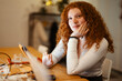 © Studio Marmellata - A young woman with curly red hair uses a tablet at a wooden table, holding a pen and looking towards the camera.