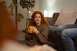 © Studio Marmellata - A young woman with curly red hair sits on the floor holding a yellow mug, smiling at the camera while a person in jeans relaxes on a sofa nearby.