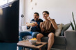© kleberpicui - Father and teenage son cheering together while watching a football match at home. Happy sports fans sitting on the sofa with snacks and drinks in a cozy living room.