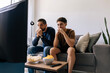 © kleberpicui - Father and teenage son nervously watching a football match at home. Emotional sports fans reacting with tension while sitting together on the sofa with snacks and drinks.