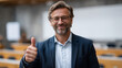 © Maksym - University administrator posing with a thumbs-up sign in a lecture hall, warm professional smile, blurred whiteboard and seating behind, balanced composition, modern academic inter