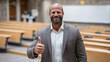 © Maksym - University director posing with a thumbs-up in a modern lecture room, friendly smile directed at the camera, rows of seats and presentation board in the background, clean education