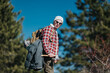 © qunica.com - A hiker wearing a red plaid shirt, white beanie, and sunglasses carries a large backpack with gear. He stands in a forested area with a clear blue sky, ready for a nature trek.