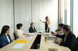 © supAVADEE BUTRADEE - female employee shakes hands with a new male employee who is introduced to her by his male CEO. Behind them, a group of fellow employees are chatting in a seminar room.
