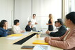 © supAVADEE BUTRADEE - female employee shakes hands with a new male employee who is introduced to her by his male CEO. Behind them, a group of fellow employees are chatting in a seminar room.
