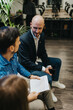 © qunica.com - One man in a blazer talks with a colleague while holding a smartphone. The scene shows a modern office with a conference table, plants, and paperwork.