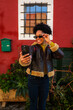 © DusanJelicic - Young woman smiling and adjusting her sunglasses while taking a selfie with her smartphone against a vibrant red wall with a window and plants, enjoying a moment of travel and connection