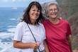 © luciano - Couple of happy elderly caucasian women standing at sea looking at camera smiling. Two elderly happy females enjoying nature and freedom