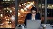 © MUGISHA - Professional businesswoman working late hours on laptop in modern office with city lights bokeh background. Dedicated female executive focus concentration overtime