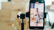 © tirachard - Close up youth Asia lady owner small business online shop sit front of phone look at camera live streaming talk to customer order promotion with full carton box at home. Shipping and delivery concept.