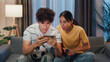 © tirachard - Asian new marriage couple sit on sofa excited cheering watching football sport game on mobile phone in living room at home. Young couple sport fans hold football watching online score on smartphone.