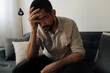 © kleberpicui - Adult man sitting on a sofa with his hand on his head showing sadness and emotional fatigue. Concepts of depression, anxiety and mental health struggles