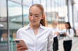 © Iryna - Young woman checks her phone while colleagues work in the background on a busy day at the office