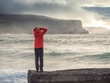 © mark_gusev - Tourist in red jacket is standing on a small rock with spectacular nature scenery with cliff, ocean and dramatic sky in the background. Doolin, area, Ireland. Travel and tourism.