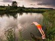 © mark_gusev - A man is jumping into a pond wearing an orange swimsuit at dusk. The sky is cloudy and the water is calm, colors are muted and calm. Sport and relaxation after a long working day concept.