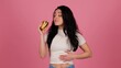 © Vlad - Smiling woman holding burger and going to bite it while rubbing her belly with her hand, isolated on pink background. Unhealthy food concept. Real time