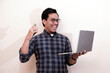 © Nurma - Southeast Asian man wearing flannel shirt standing and smiling expression happy with holding laptop and mobile phone, on white isolated background.