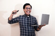 © Nurma - Southeast Asian man wearing flannel shirt standing and smiling expression happy with holding laptop and mobile phone, on white isolated background.