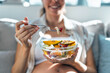 © nenetus - Pregnant woman eating a bowl of fruit salad while sitting on the couch at home