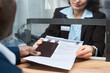 © pressmaster - Caucasian middle aged woman processing visa application documents for Black man at service counter, woman handing over passport and paperwork with identification photos visible on desk