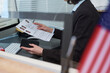 © pressmaster - Middle aged Caucasian woman processing visa applications at visa center, holding passport and documents with applicant photos, sitting at desk with computer, American flag visible in foreground