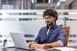 © Tetiana - Concentrated young Indian man wearing glasses sitting at a desk in the office and working on a laptop