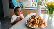© velikiyzayats - Excited young Black boy wearing bunny ears looking at a plate of traditional hot cross buns in a kitchen.