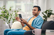 © dodotone - Indian man using smartphone relax sitting on sofa in living room
