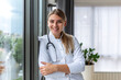© Graphicroyalty - Portrait of smiling female doctor standing by window. Confident medical professional with stethoscope. Healthcare and medicine concept. Successful young woman practitioner in white coat.