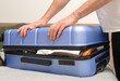 © valiantsin - Man pressing blue suitcase lid to close luggage. Male securing packed case on bed surface. Travel organization, baggage control, departure readiness concept..