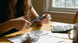© Stock x - A focused woman sits at a sunlit wooden table cluttered with receipts, bills, and a calculator, cutting a document with scissors to organize expenses and plan a personal budget