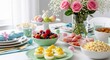 © Rana Graphic - Festive Easter brunch spread with deviled eggs, sliced ham, fresh berries, and pastel eggs. Pink roses in vase and green herbs garnish food. Table set for spring holiday meal celebration with guests.
