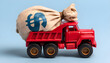 © Oleksiy - A red toy dump truck carries a large beige money bag marked with a dollar sign against a blue background. The scene evokes themes of wealth, transportation, and savings in a playful manner