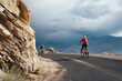 © Soloviova Liudmyla - Couple of mountain bikers riding on asphalt road near massive jagged cliff in Leh, Ladakh India featuring white monastery on distant hill under dramatic dark stormy sky in Himalayan mountain desert