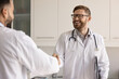 © fizkes - Friendly medical partnership. Smiling young male physician in white coat hold clipboard under arm exchange handshake with doctor colleague after successful meeting express gratitude for advice support