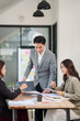 © aekachai - A group of three young Asian business colleagues—two women and one man—engaged in a productive meeting. They are leaning over a wooden table, reviewing documents, taking notes, and discussing strategi